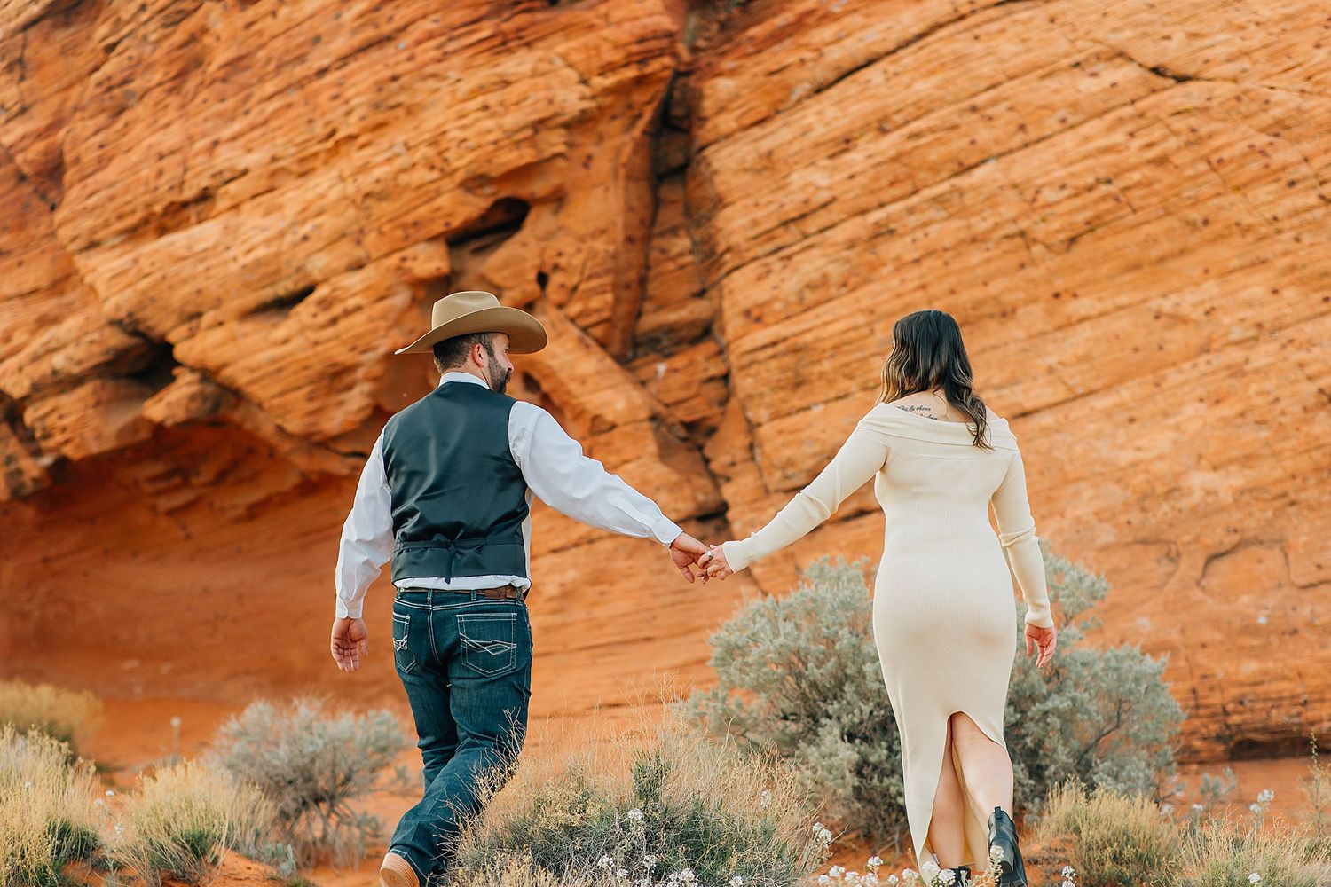Couple holding hands while walking along scenic red rock canyon walls in desert landscape.