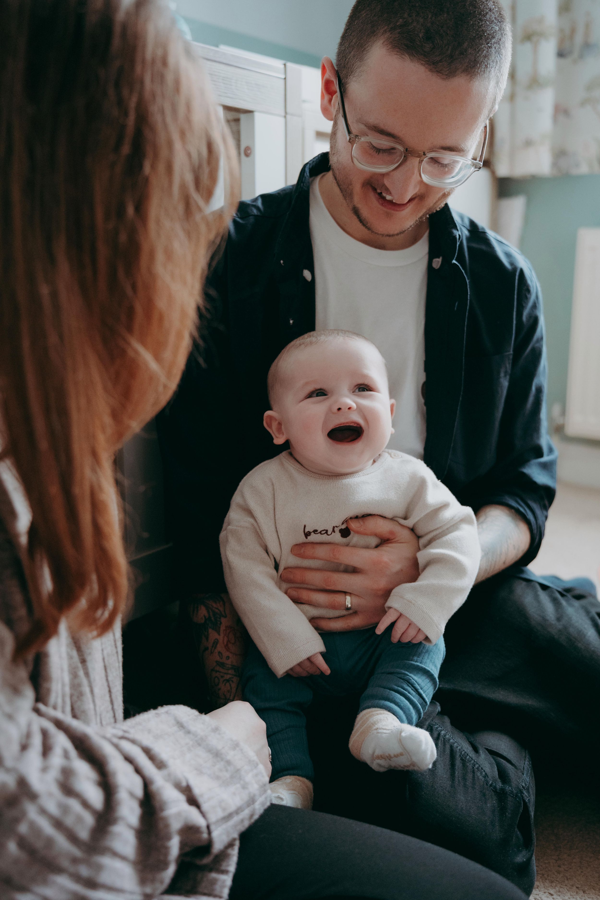 A baby in a white sweater laughs joyfully while being held during an intimate indoor family photoshoot.