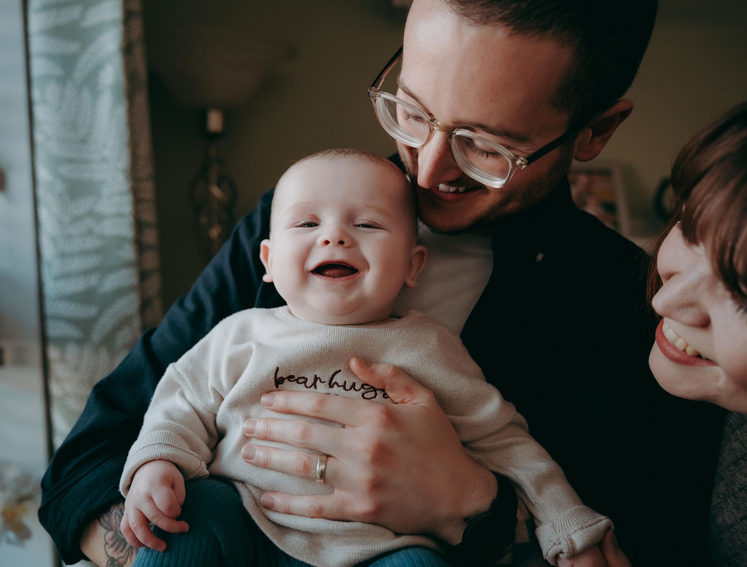 A happy baby in a white onesie smiles while being held by adults in a cozy indoor setting.