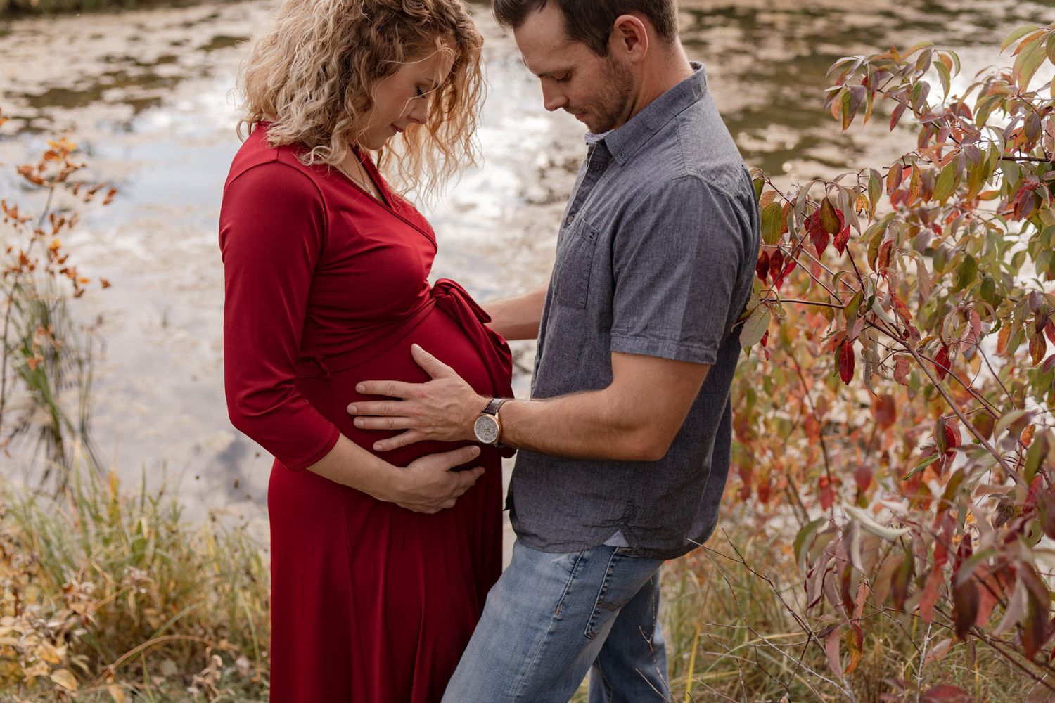 maternity couple hold baby belly while standing among fall foliage at cataraqui creek conservation area, kingston ontario
