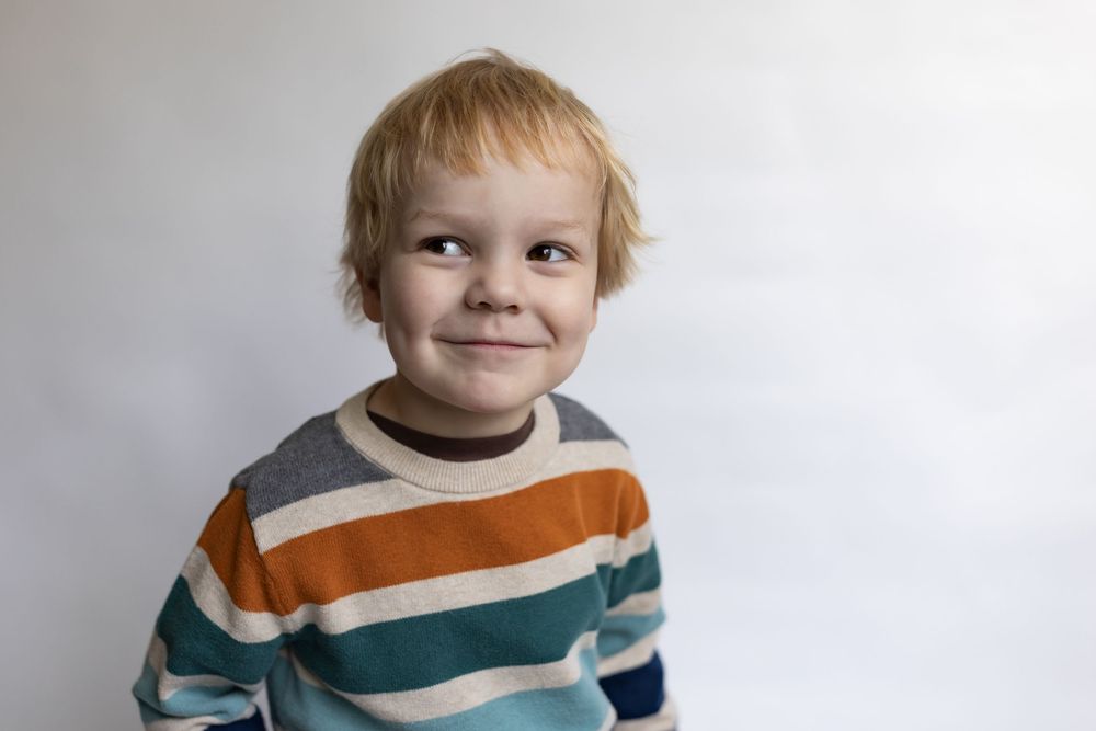 Cute young boy with blonde hair giving a sly smile, wearing a striped sweater against a white background.