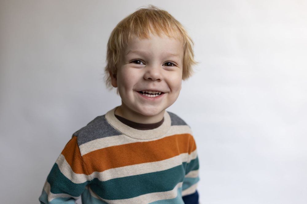 A happy toddler boy with blonde hair grins widely in a colorful striped sweater.