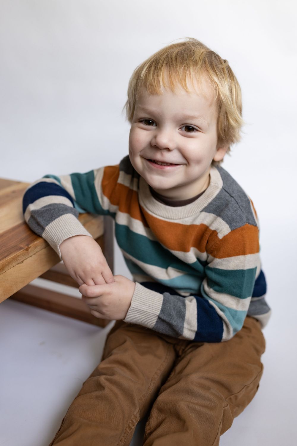 A cheerful toddler boy in a striped sweater sits on a wooden bench, smiling at the camera.