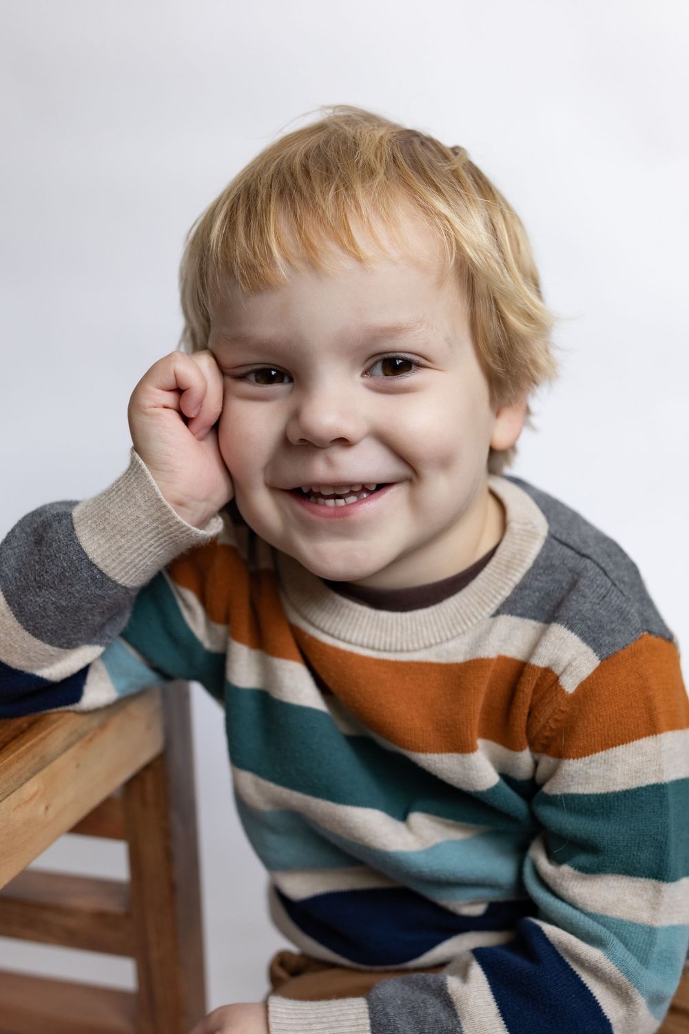 A laughing toddler boy in a multicolored striped sweater rests his hand on his cheek.