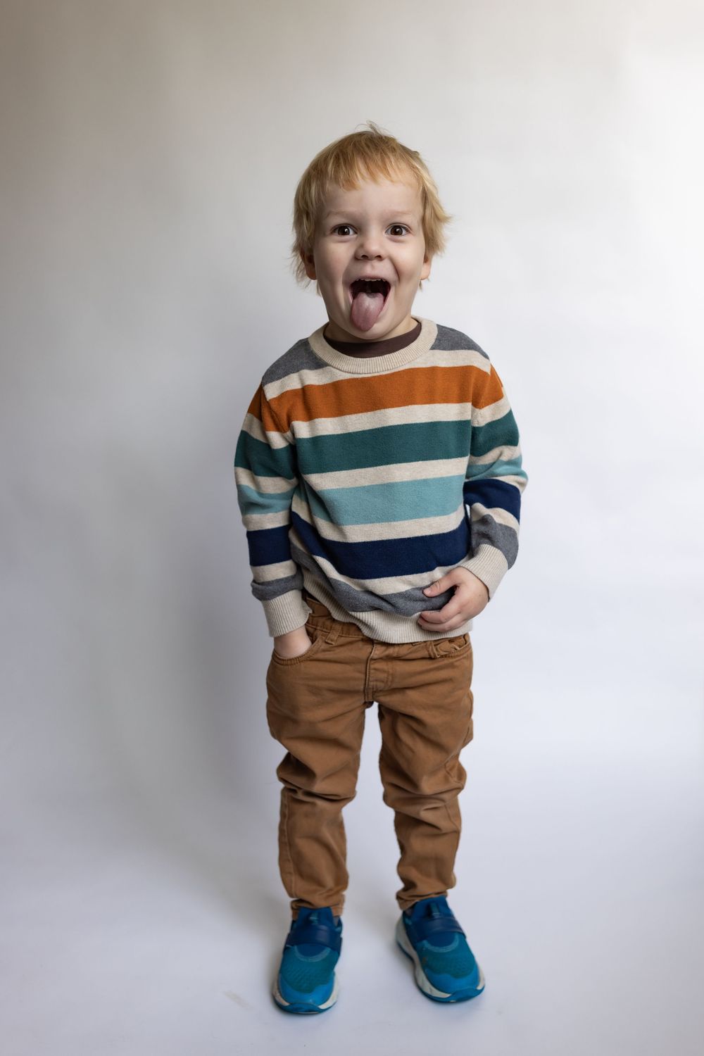 Playful toddler boy sticking out tongue, wearing colorful striped sweater, tan pants, and blue sneakers.