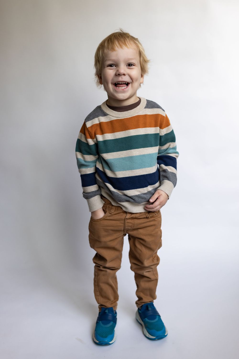 Happy toddler boy laughing with hands in pockets, wearing a multicolored striped sweater and tan pants.