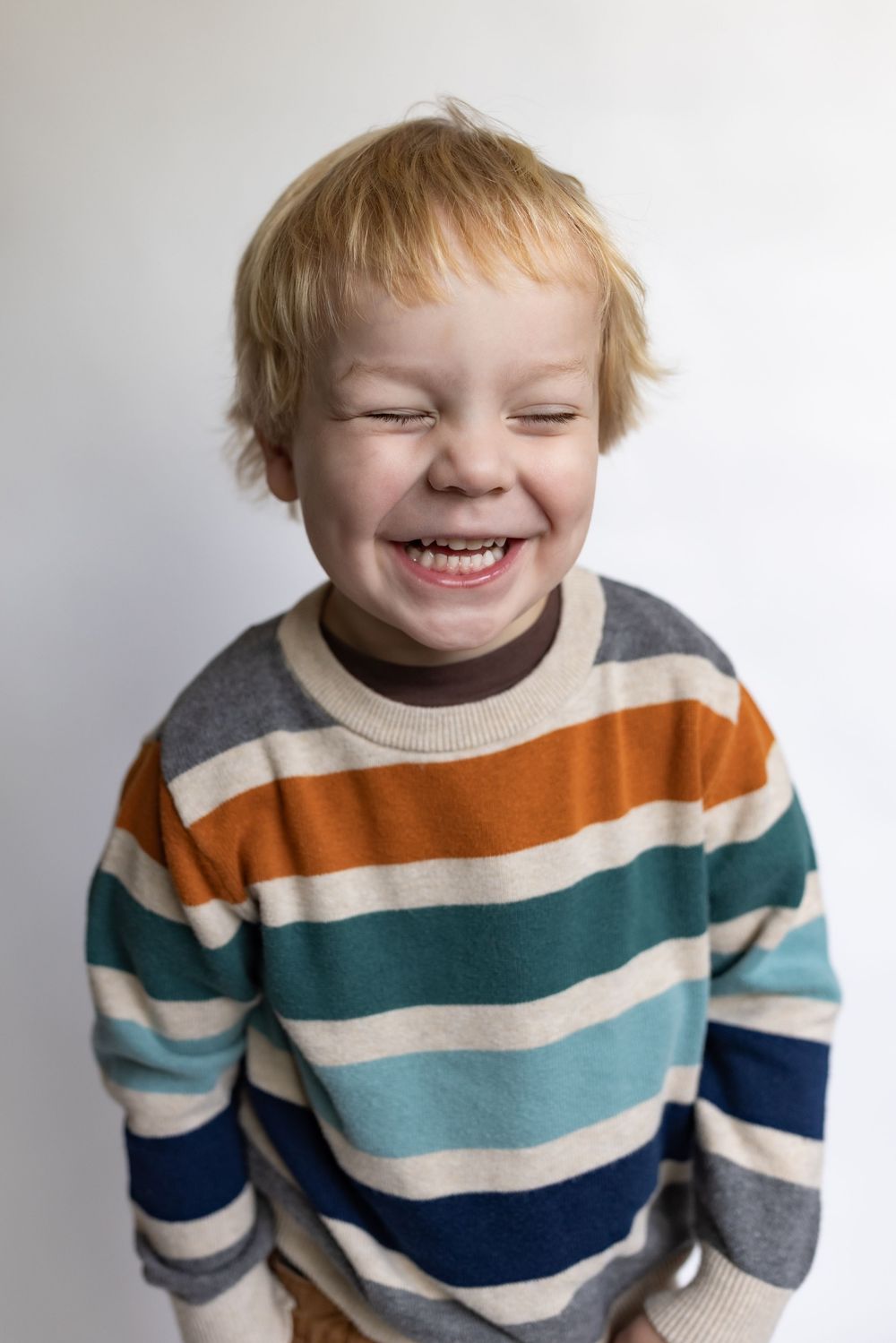 Joyful young boy laughing with eyes closed, wearing a bold striped sweater in orange, teal, and navy.