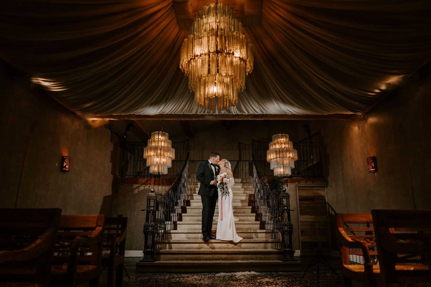 Elegant wedding ceremony venue with ornate crystal chandeliers hanging from a vaulted ceiling over rows of white chairs.