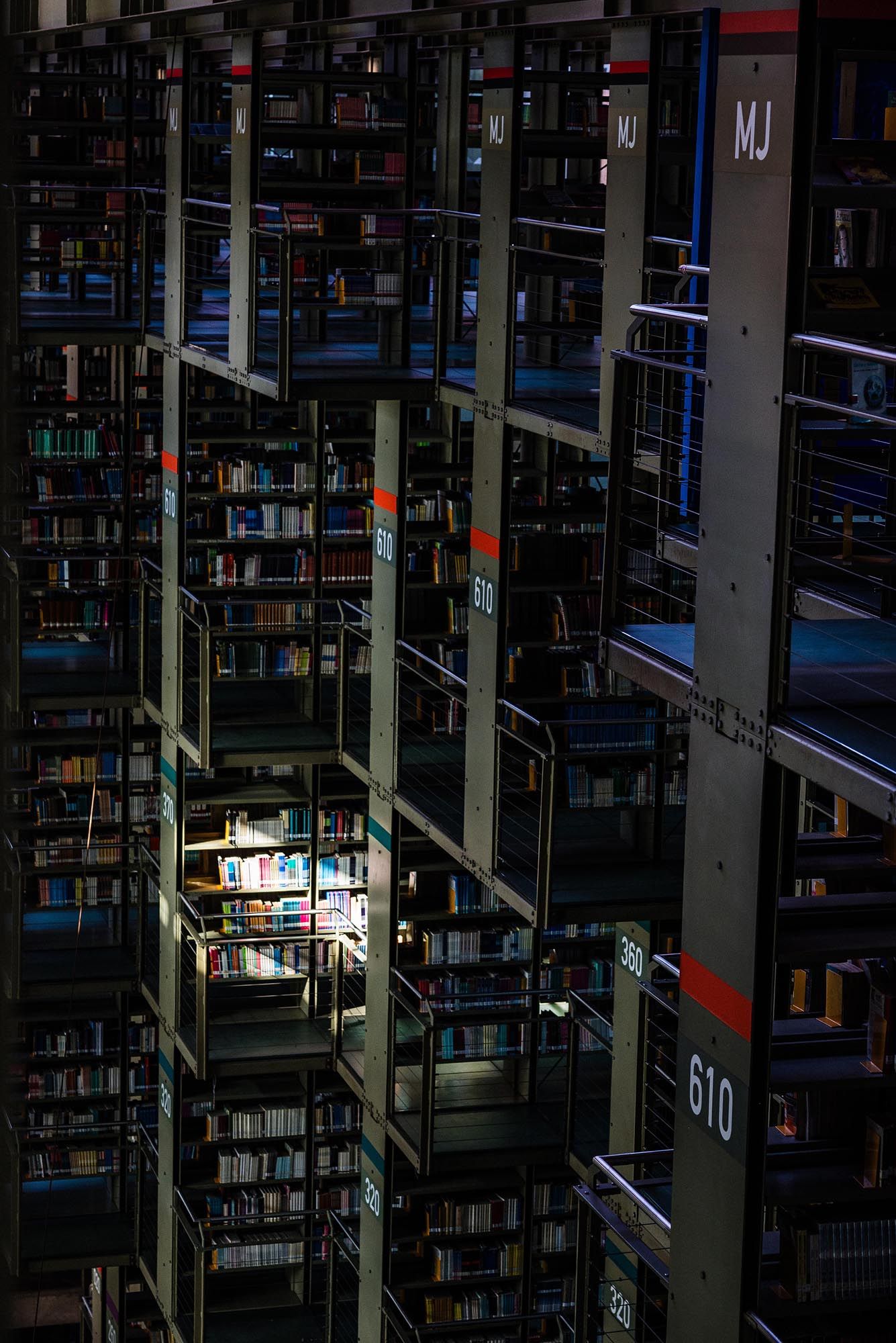 Dimly lit library shelves with glowing sections illuminated between dark stacks of books and numbered aisles.
