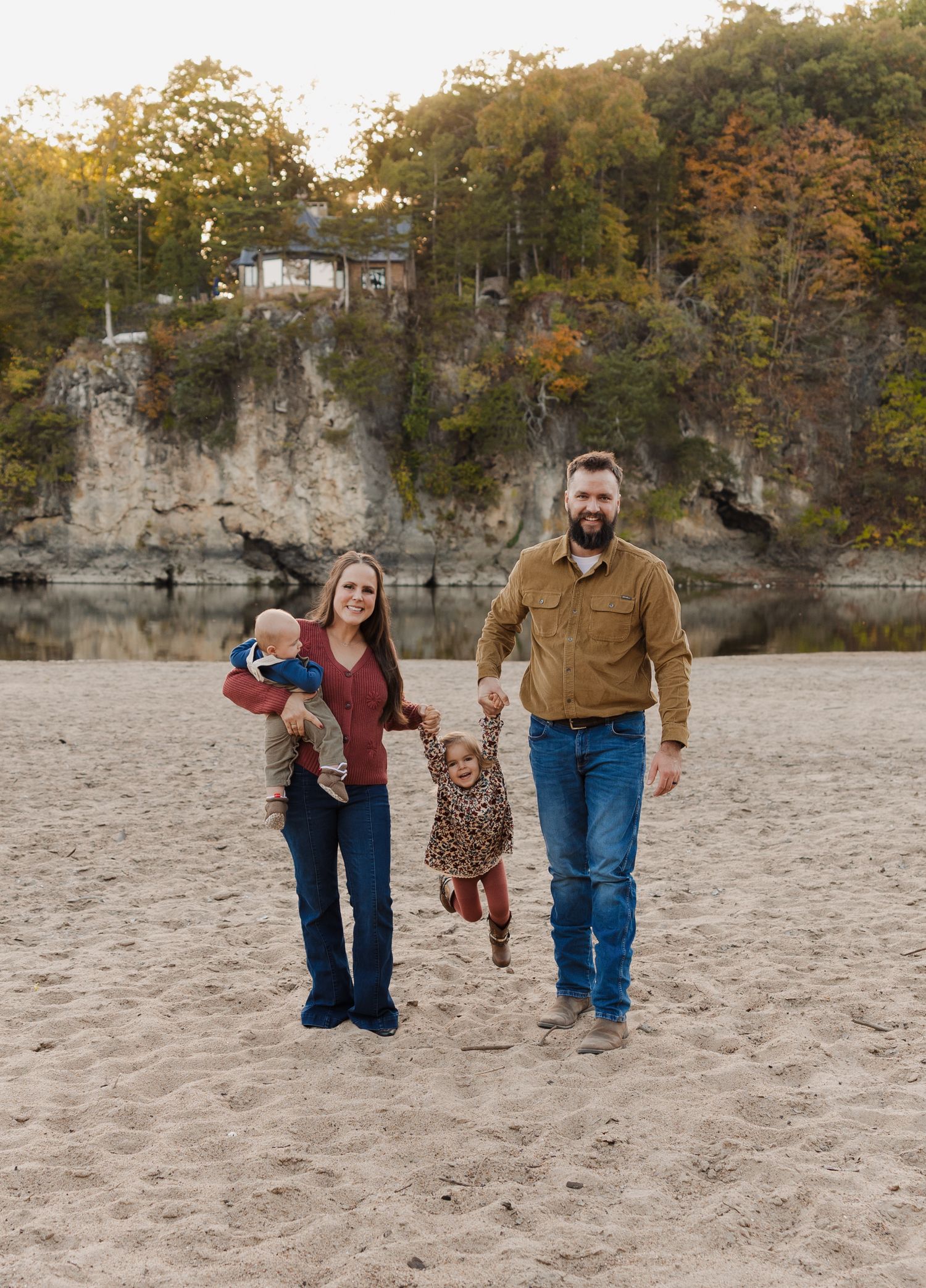 A family walks together on a sandy beach with dramatic limestone cliffs and autumn foliage in the background.