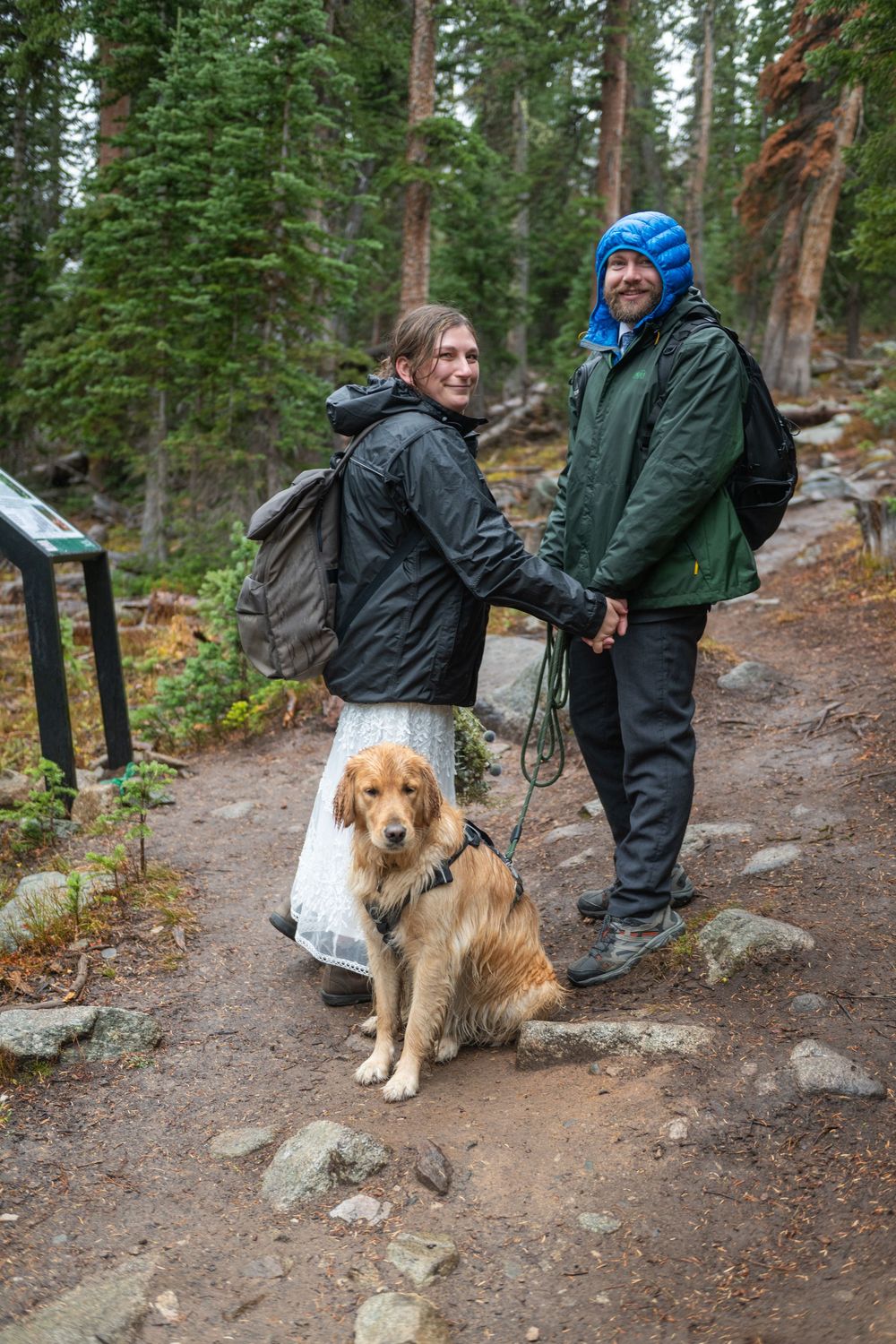 An eloping couple are hiking in wedding attire and rain coats up a mountain trail with their dog. They are facing each other, holding hands.
