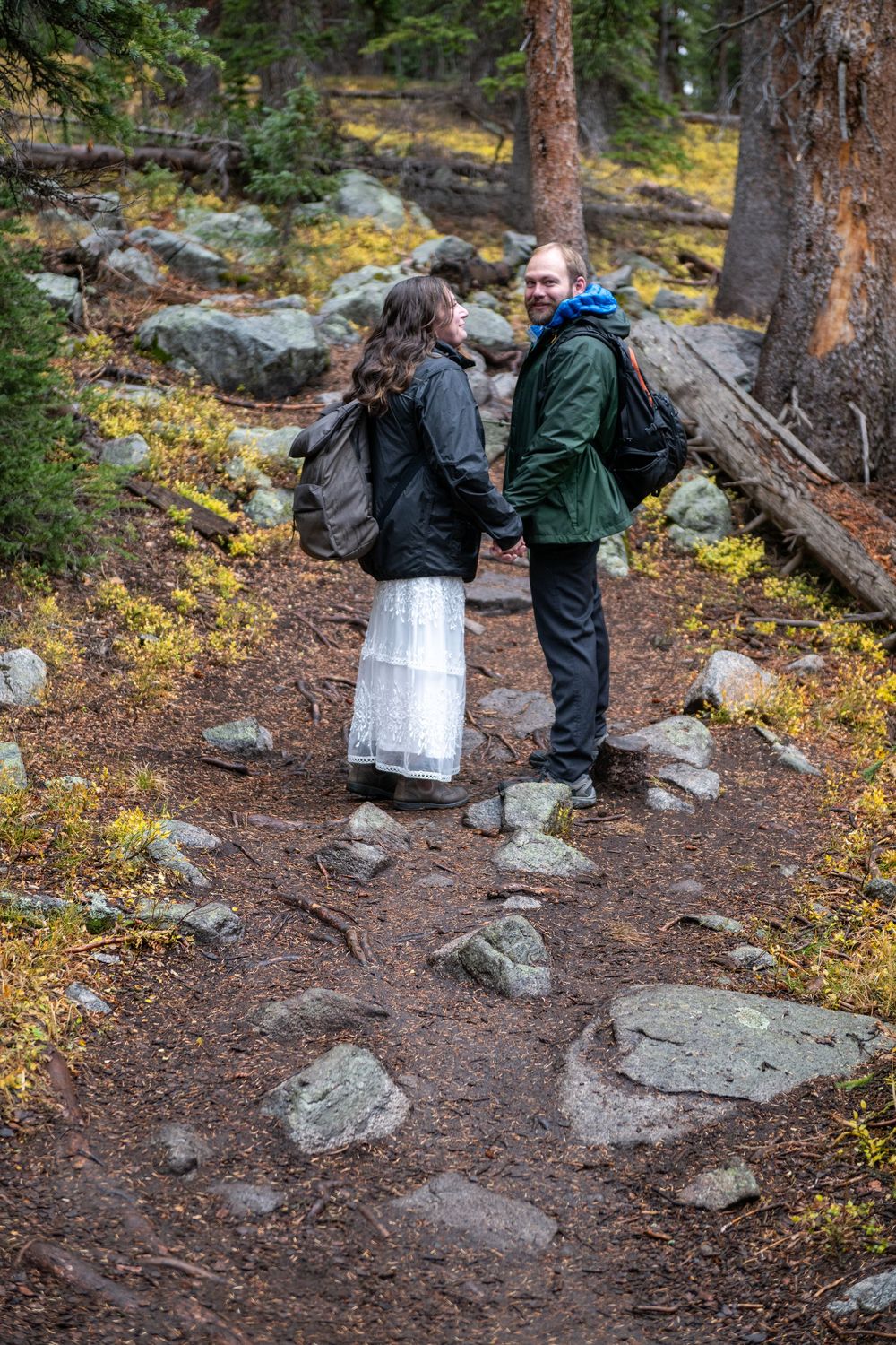 A bride and groom stand in the middle of a rocky trail facing each other, holding hands. They are wearing backpacks and puffer coats.