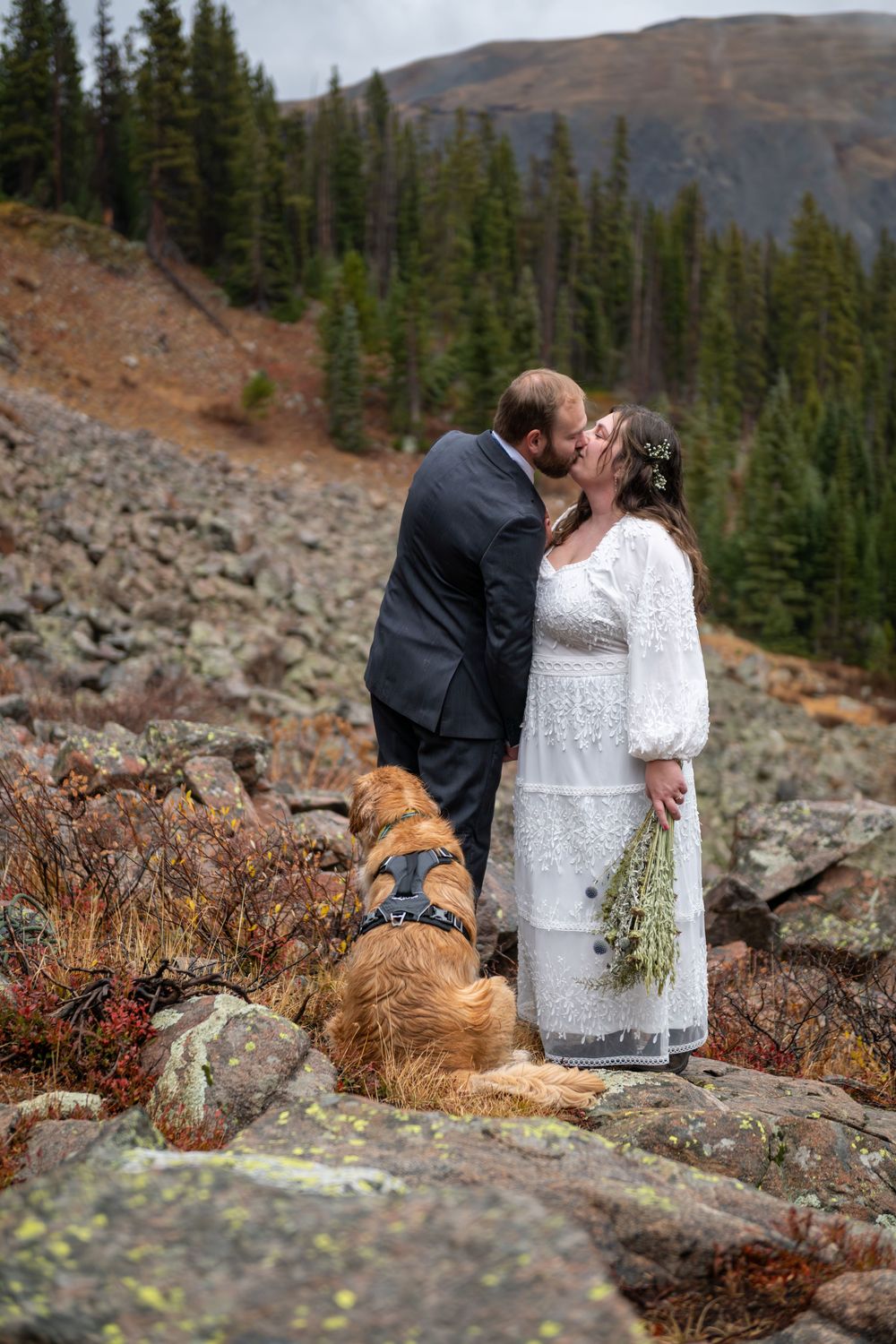 An eloping couple share a kiss,  she wears a white dress and he a suit on rocky mountain terrain with a dog and evergreen forest backdrop.
