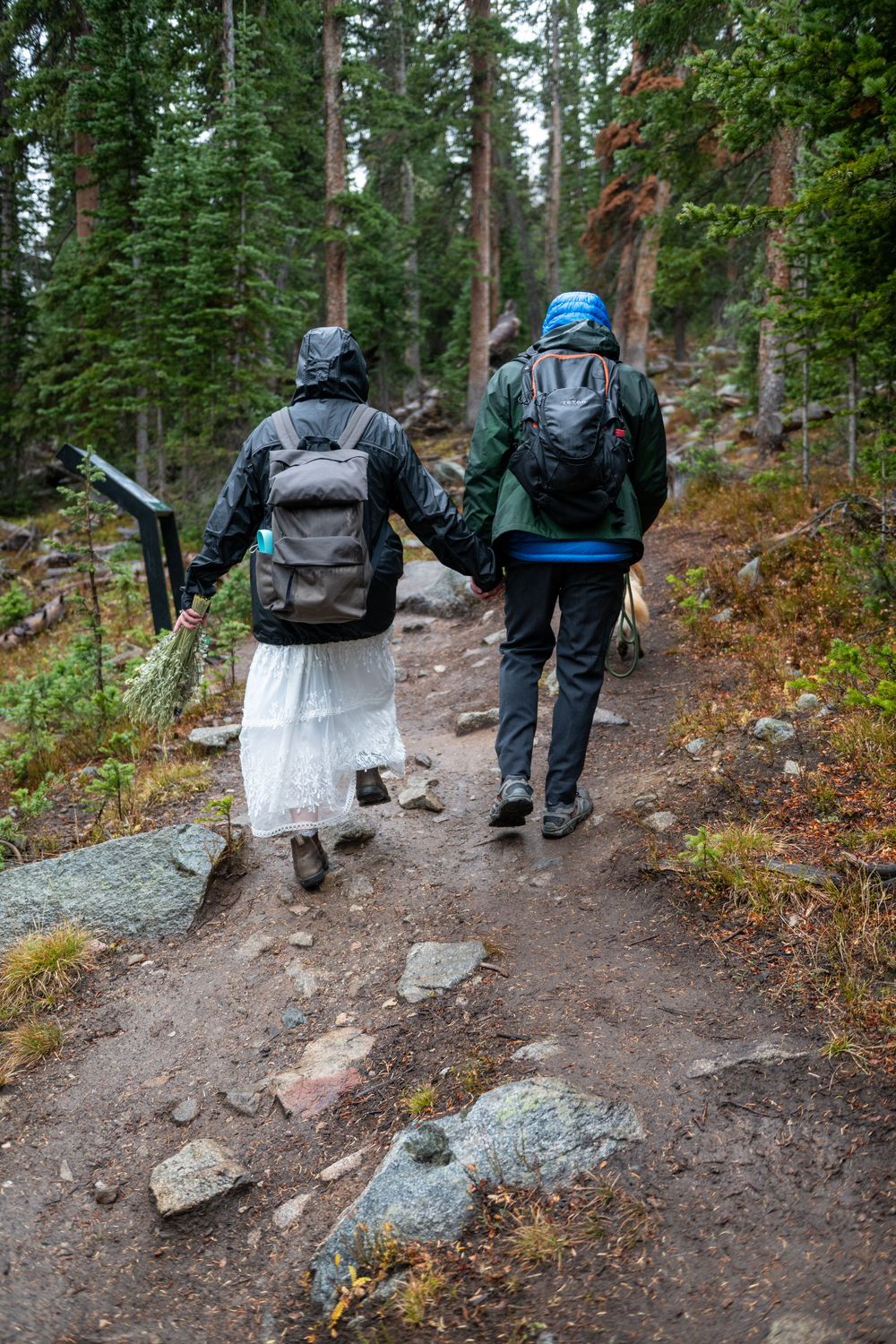 An elopign couple in wedding attire hiking a forest trail with backpacks in the rain.