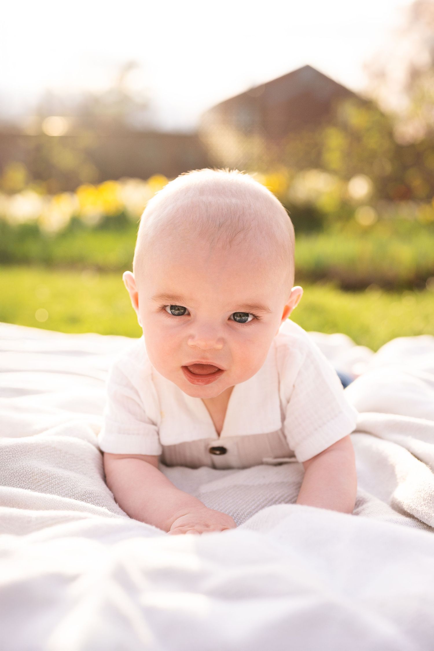 A baby in a white outfit smiles while laying on a blanket outdoors during golden hour lighting.