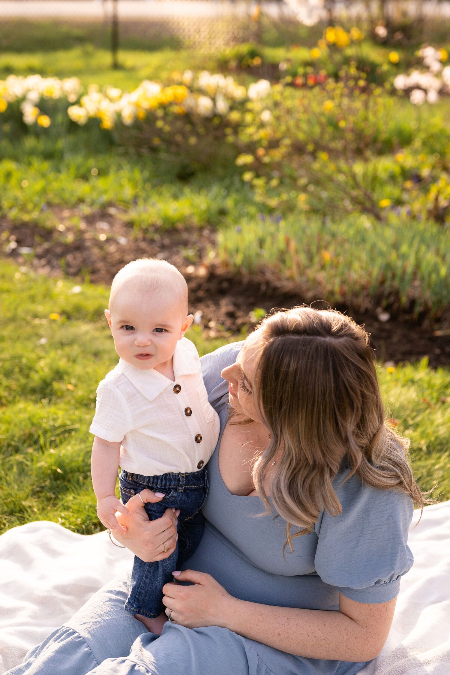 Mother and baby share tender moments on white blanket surrounded by spring flowers and greenery in outdoor setting.