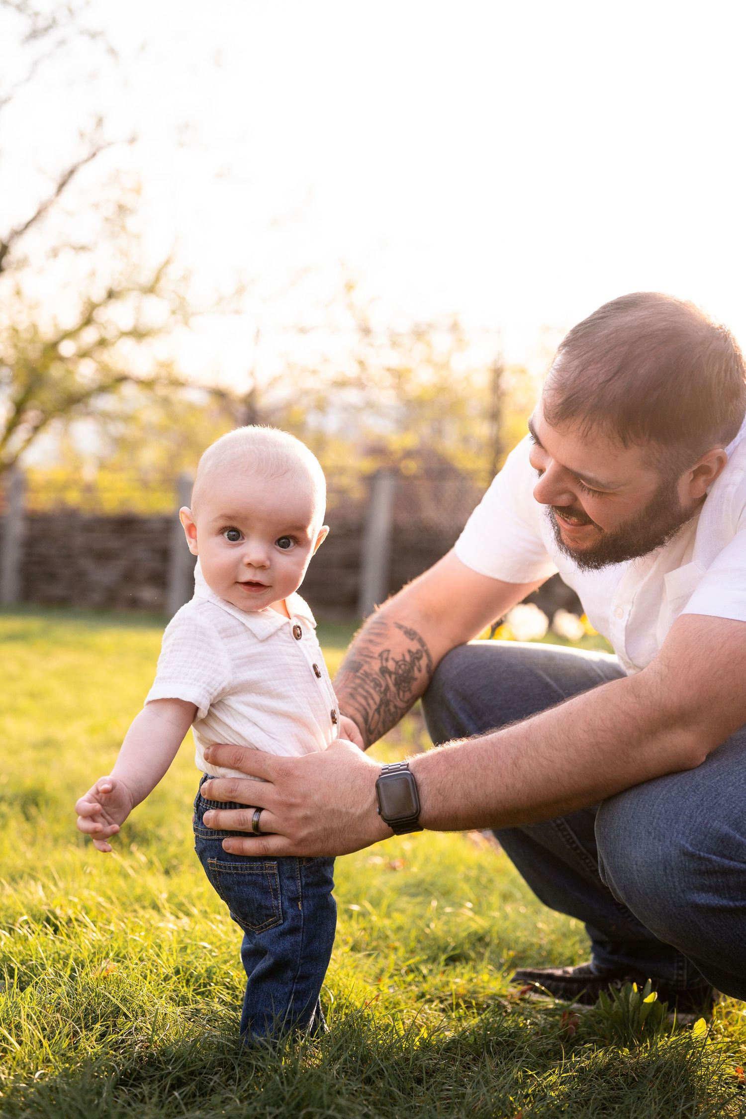 Baby learning to walk in a grassy field at sunset while supported by adult hands.