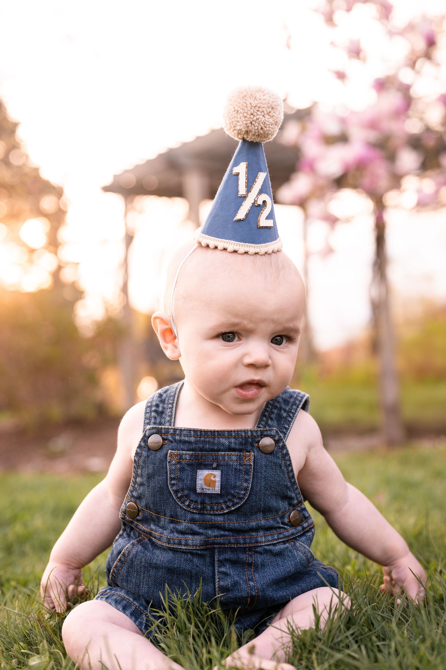 Small child wearing denim overalls and blue birthday party hat sits in grass at sunset.