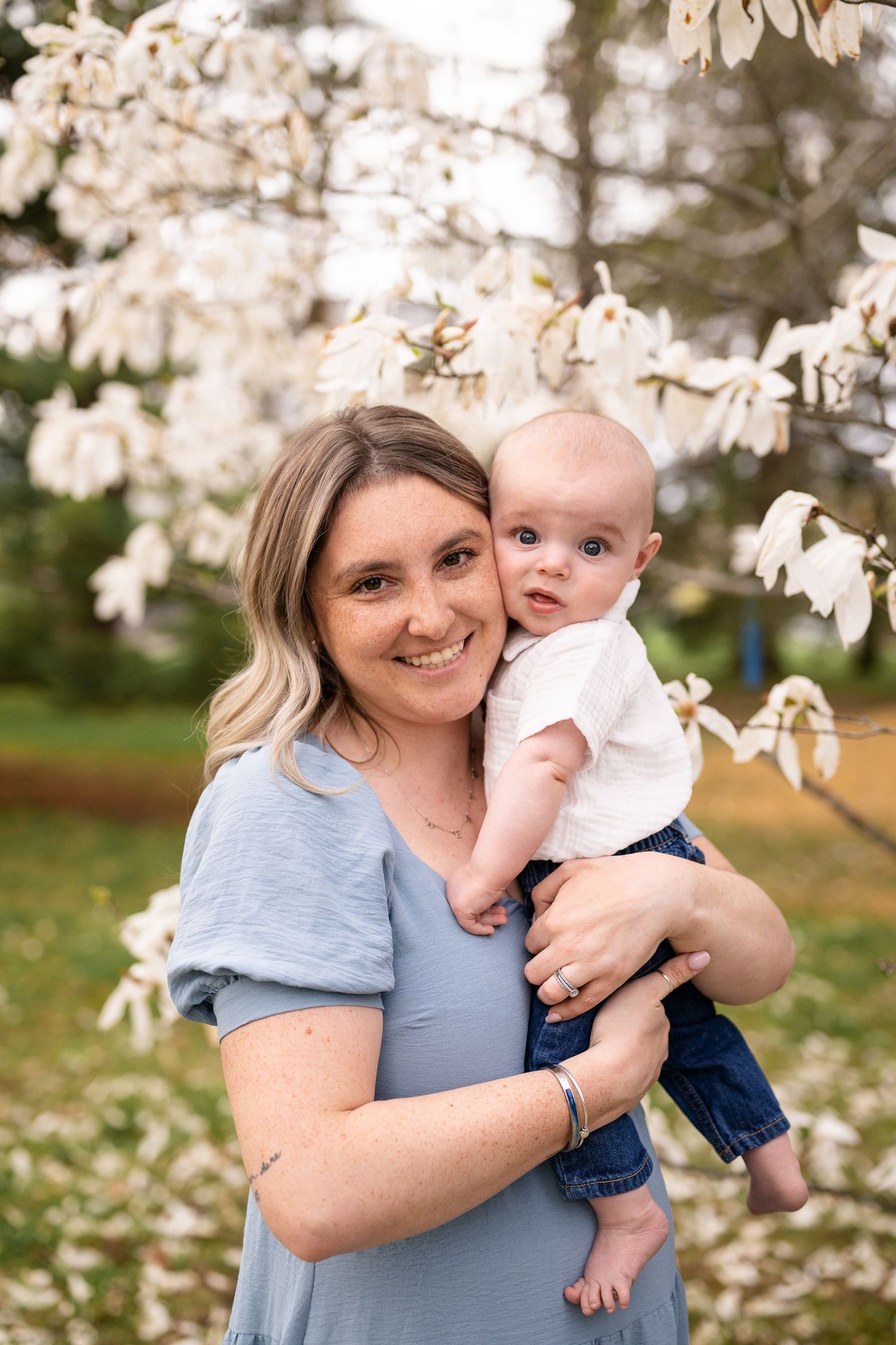 A parent and baby pose together under blooming white spring blossoms in a garden setting.