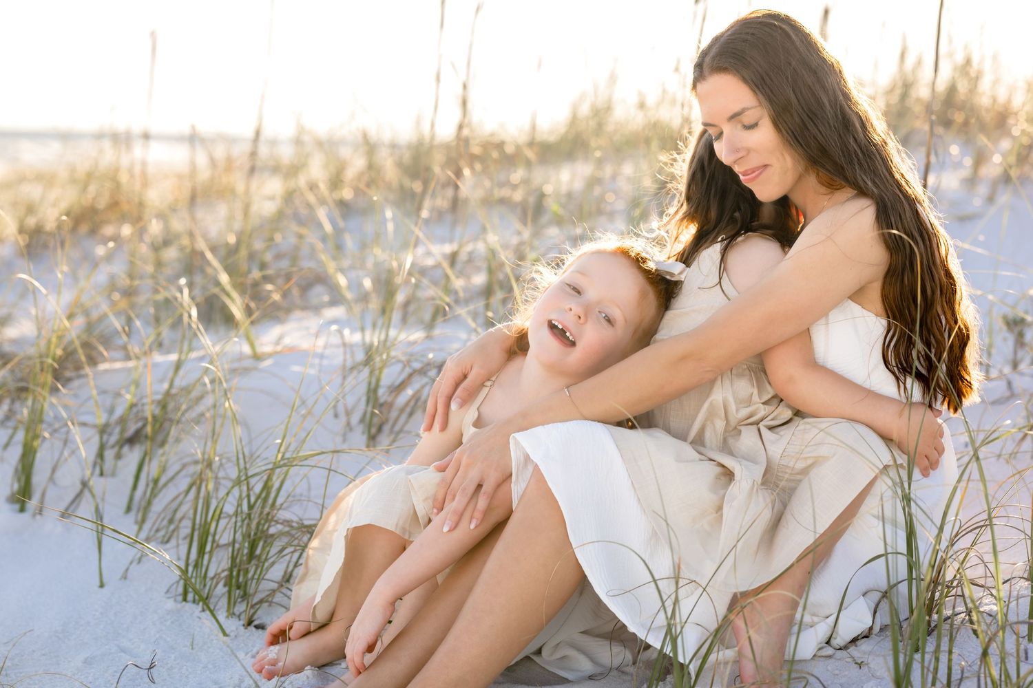 Two people in white dresses share a joyful moment while sitting together on white sand dunes at sunset.
