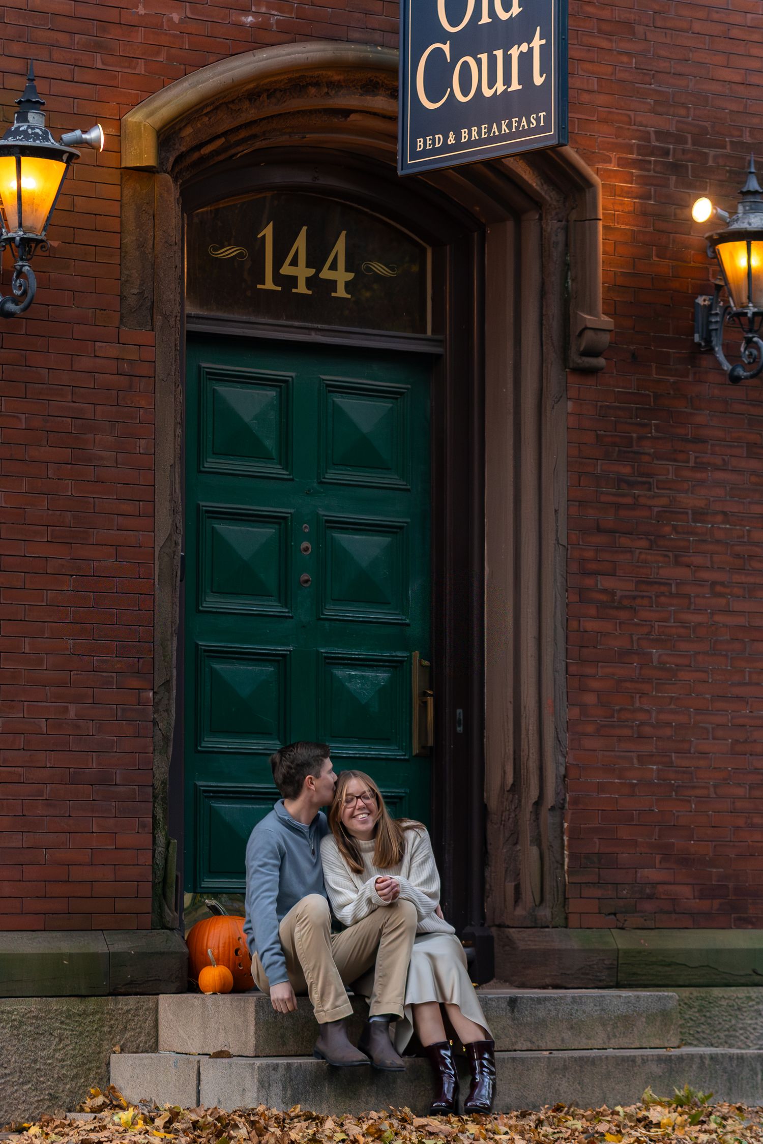 A couple sits on brick steps in front of a historic building with green doors and autumn leaves scattered nearby in Providence, Rhode Island