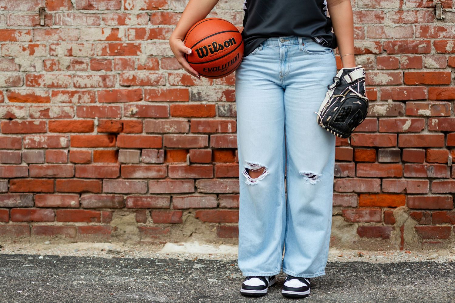 Downtown Cedar Rapids high school senior girl in light blue jeans and black shirt against brick wall holding basketball and baseball glove.
