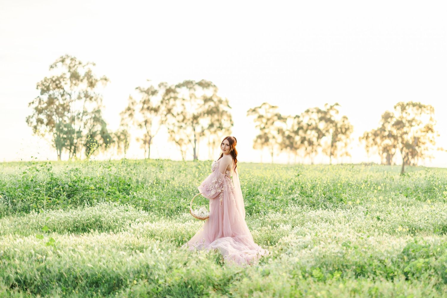 A person in a flowing pink gown walks through a sunlit meadow during golden hour creating a dreamy atmosphere.