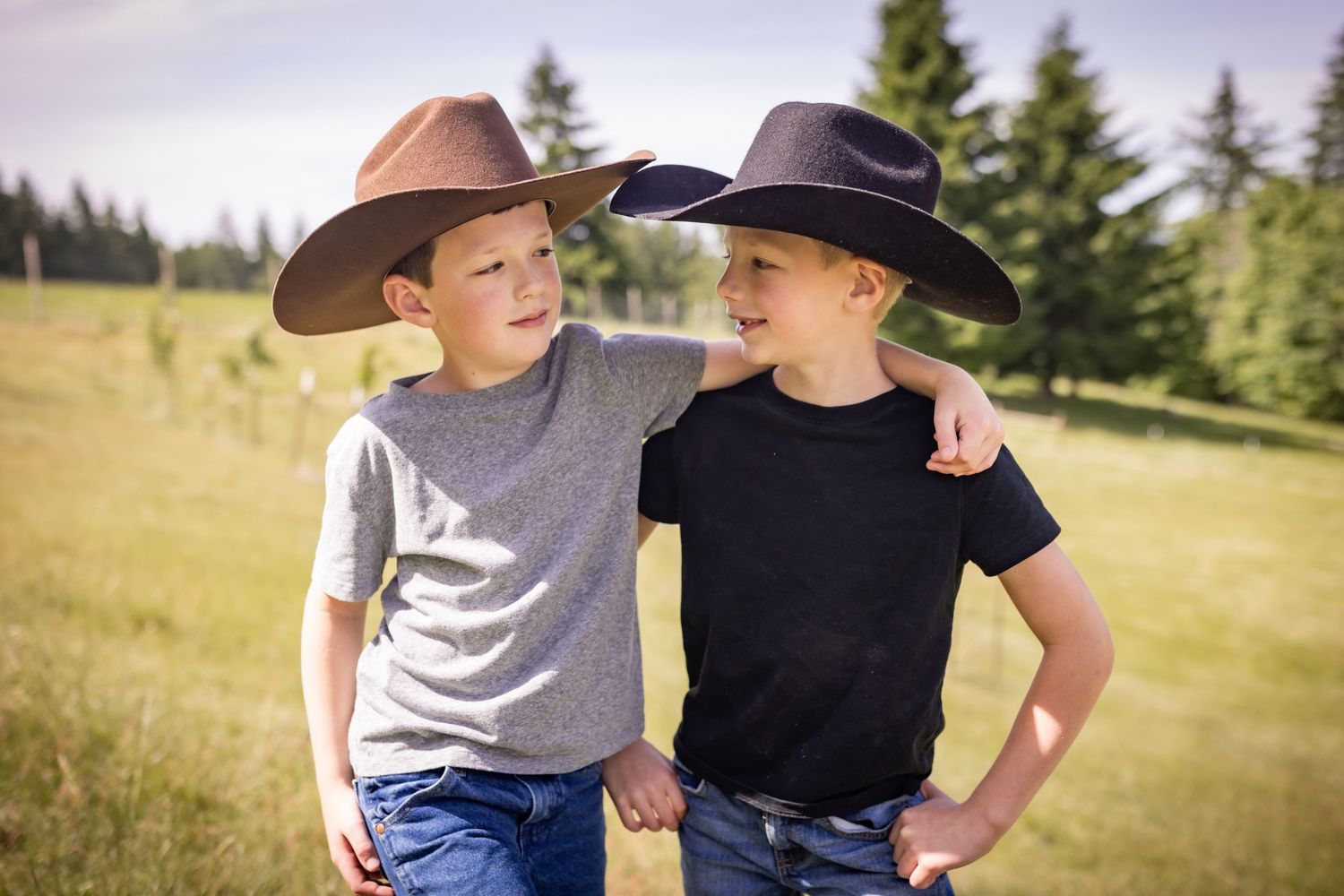 Two boys in cowboy hats smile with arms around each other in a sunny green field.