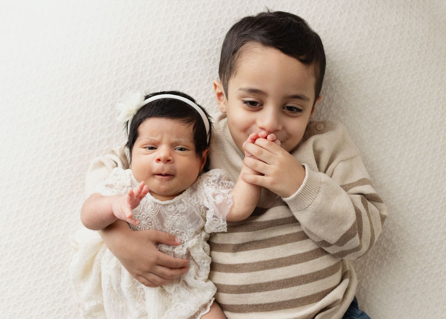 Two children lying on a cream-colored background, one wearing a lace outfit and the other in a striped sweater. Elizabeth Lang Photography S