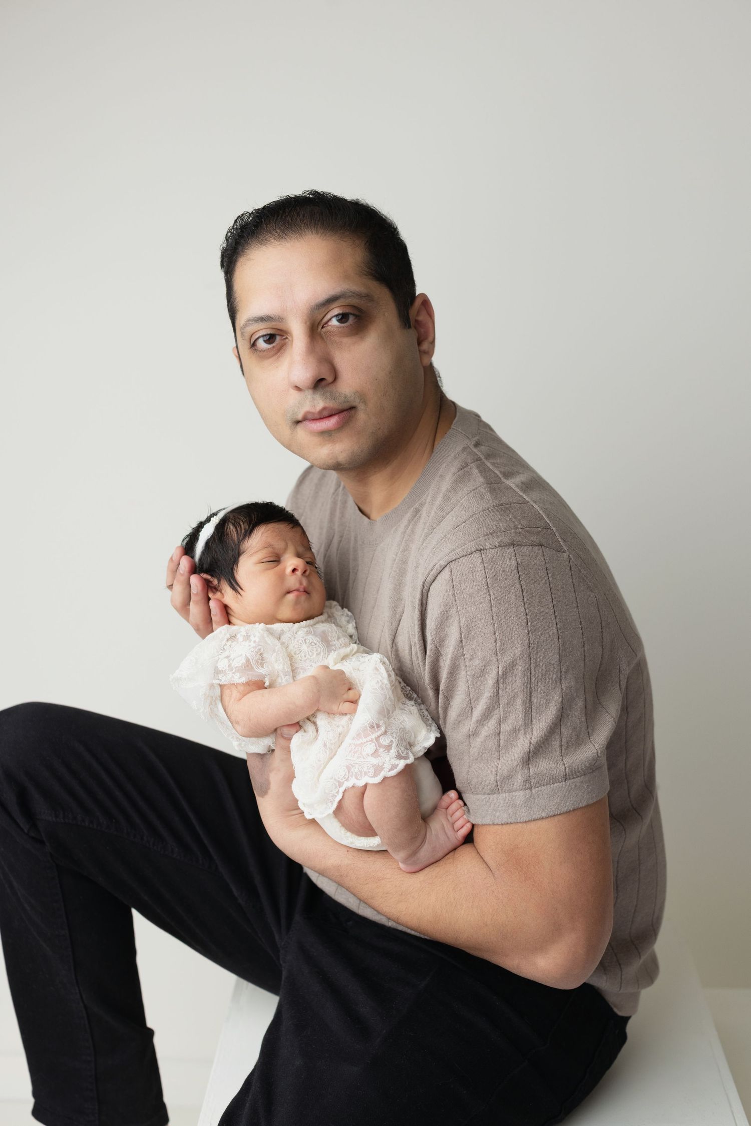 A parent in a gray t-shirt holds a sleeping newborn baby wearing white lace against a light background. Elizabeth Lang Photography Surrey