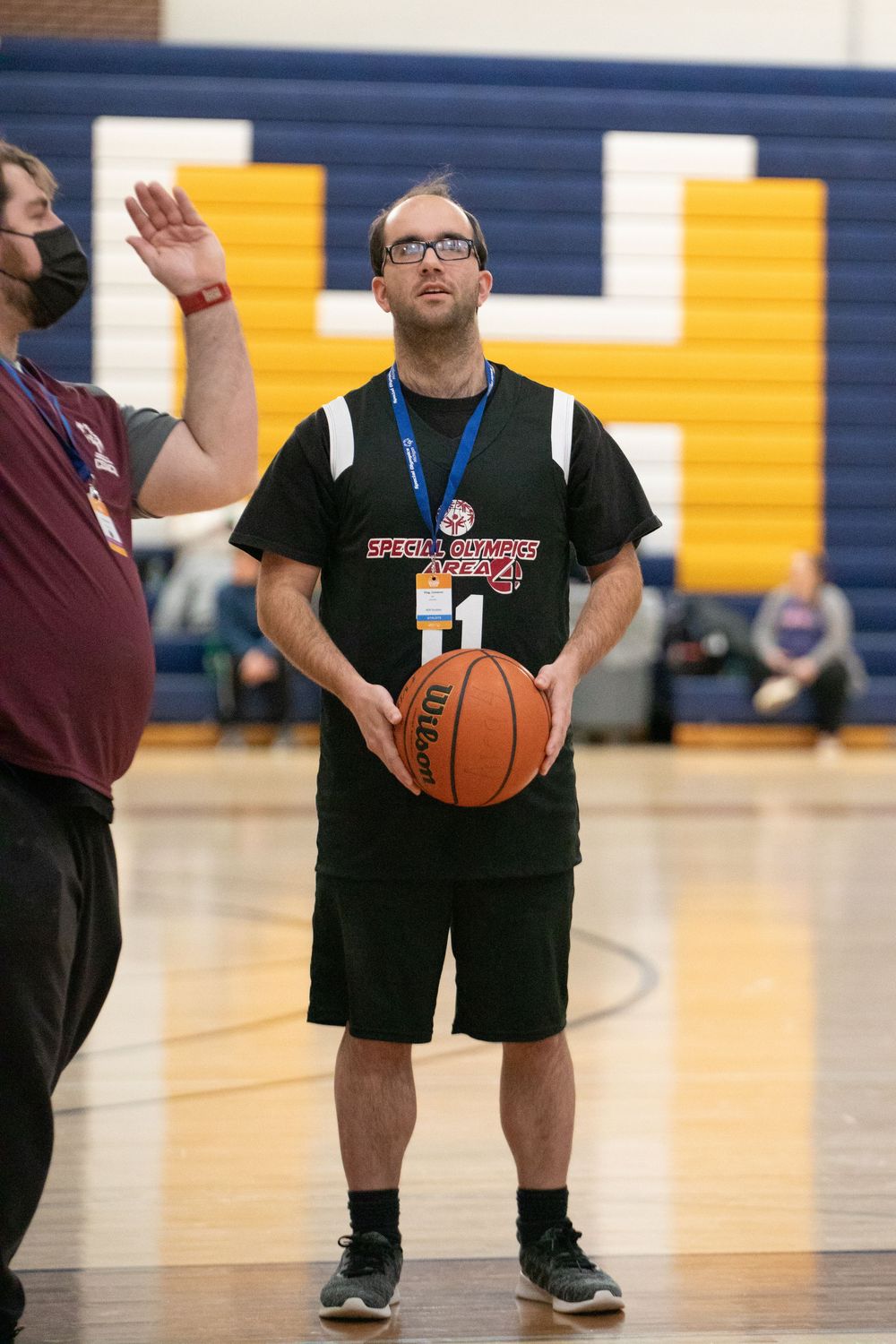A basketball player in a black uniform stands ready to shoot on a gymnasium court with yellow and blue walls.
