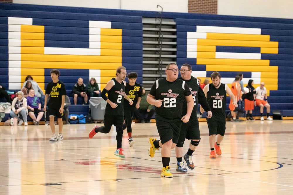 Basketball players in black uniforms run across a gymnasium court with yellow letters on the wall during a game.