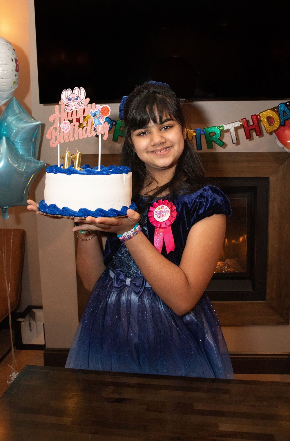 A birthday celebration features a blue and white cake with decorations and balloons against a dark background.