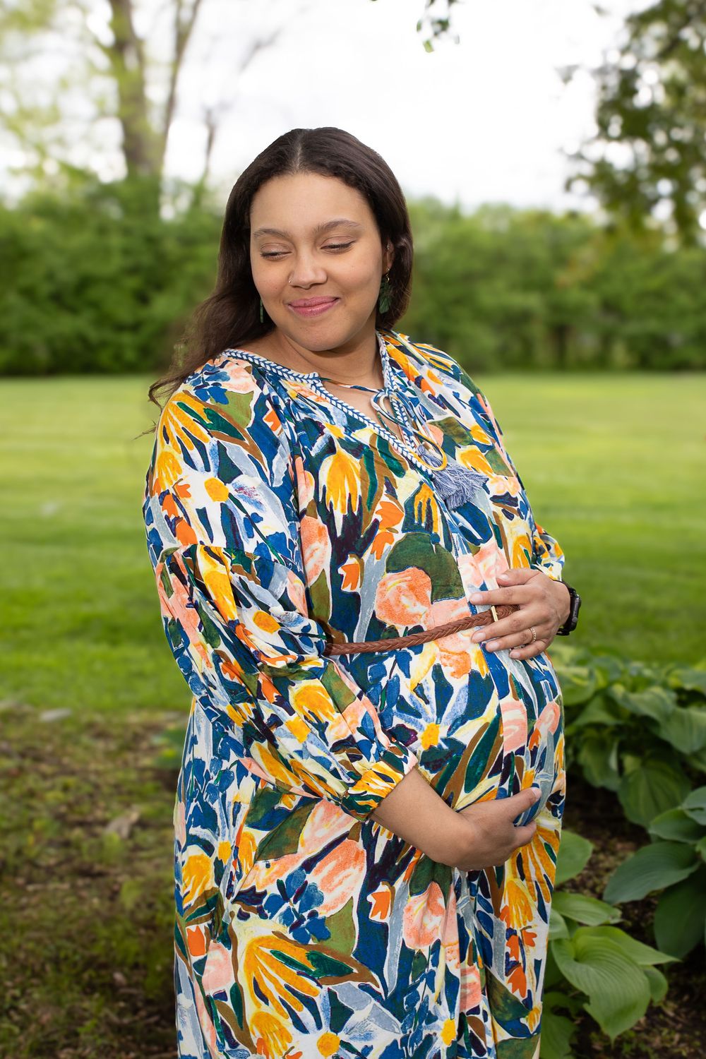 Expectant mother in colorful floral print dress stands peacefully in a lush garden setting during summer.