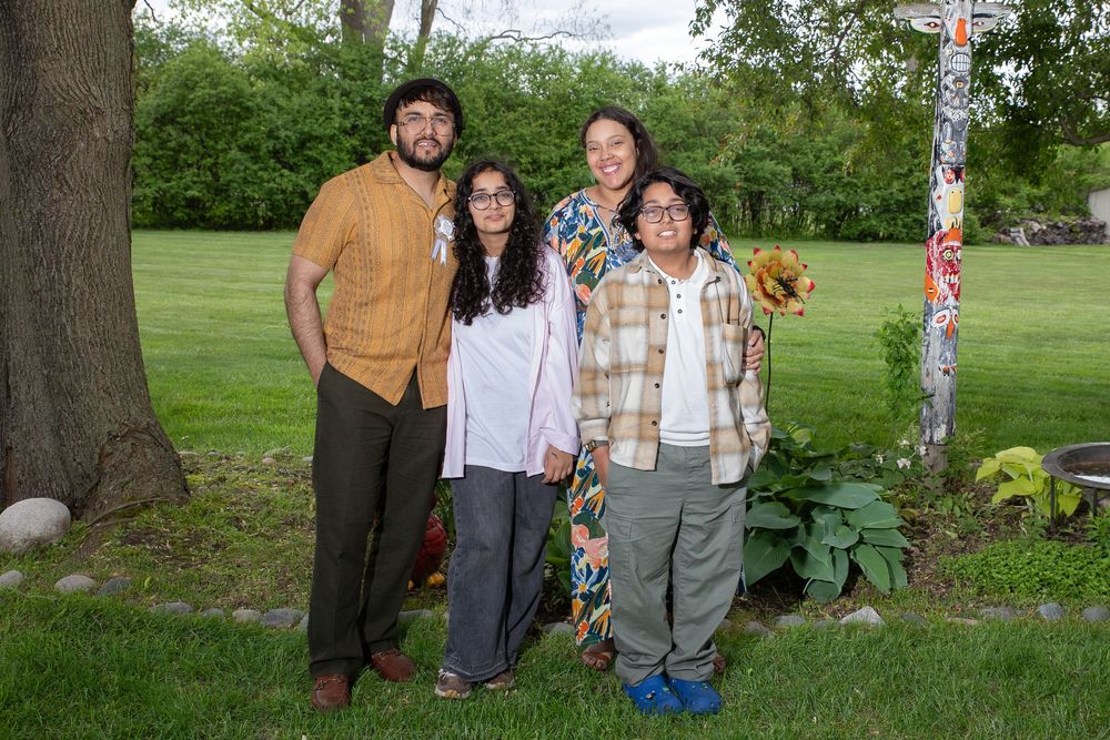 A group of people gather outdoors in a park-like setting with trees and greenery in the background.