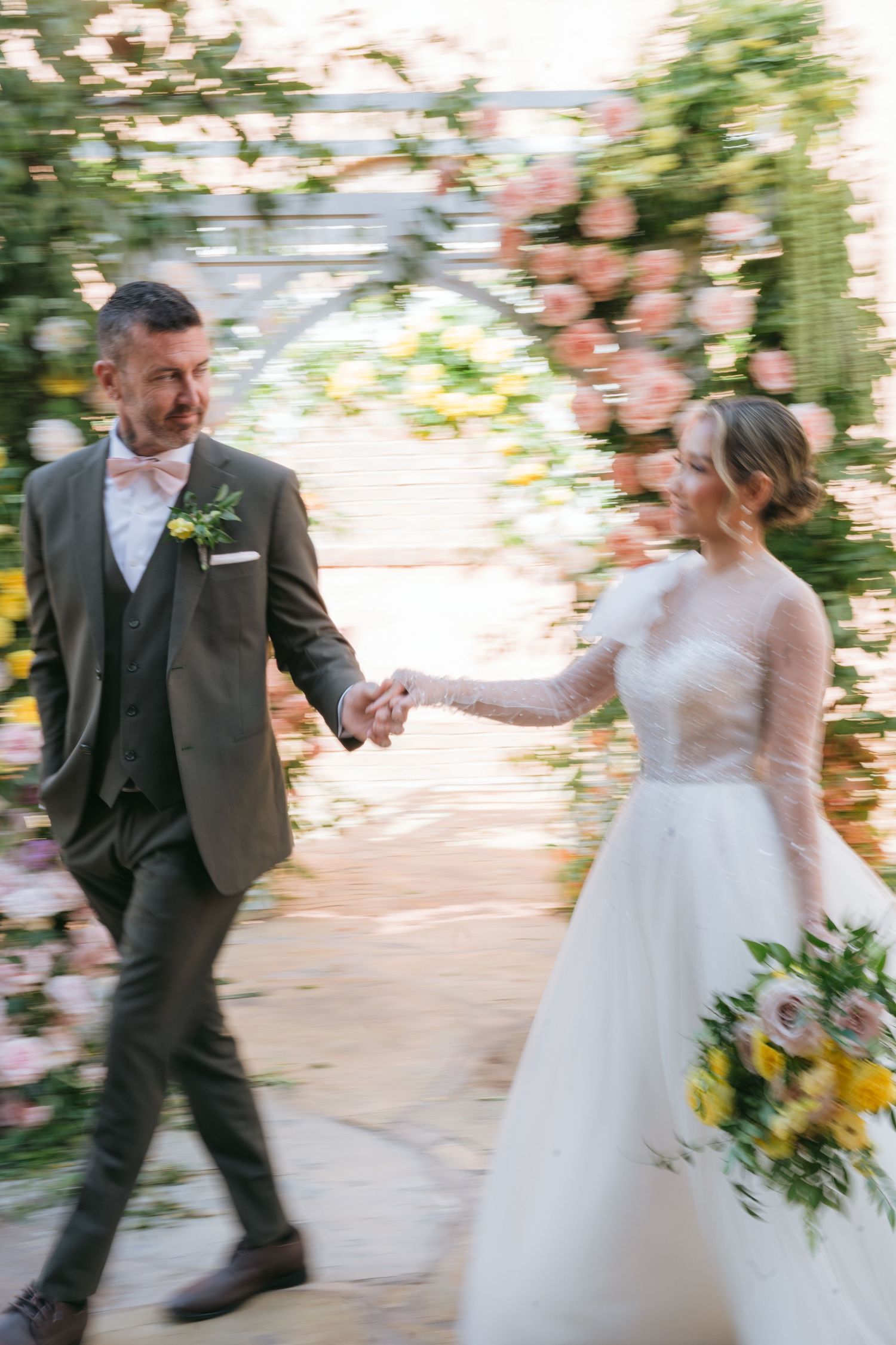 Newlywed couple dances together in a sunlit garden setting with pink floral decorations.