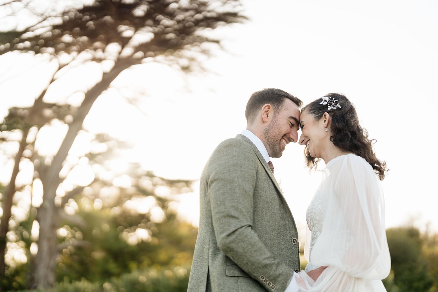 A couple in wedding attire share an intimate moment at sunset under cypress trees in a romantic outdoor setting.