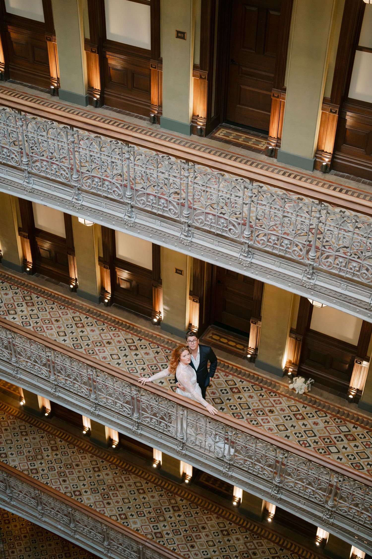 Elegant couple stands on ornate vintage staircase with intricate carved details and warm lighting in historic building interior.