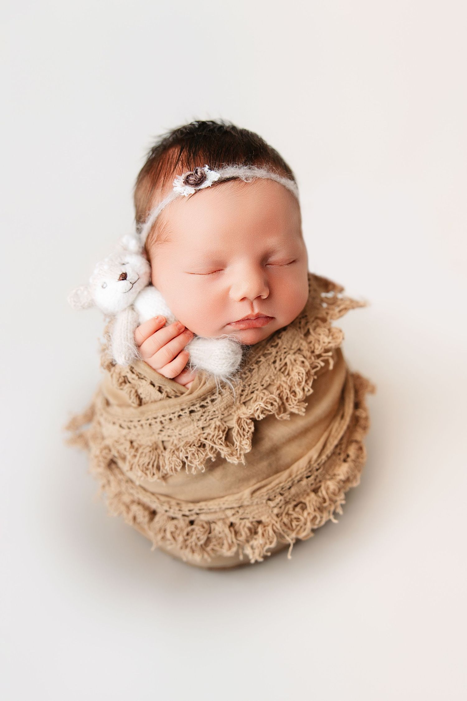 Sleeping newborn baby wrapped in soft beige lace fabric with delicate white flower headband in artistic portrait series.