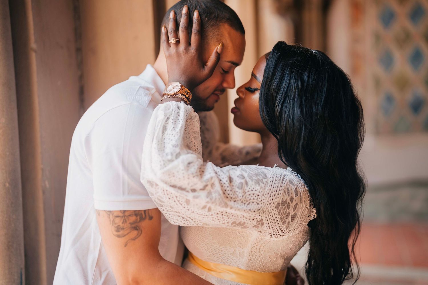 A romantic couple shares an intimate moment in front of a decorative wall, with the woman wearing a white lace dress.