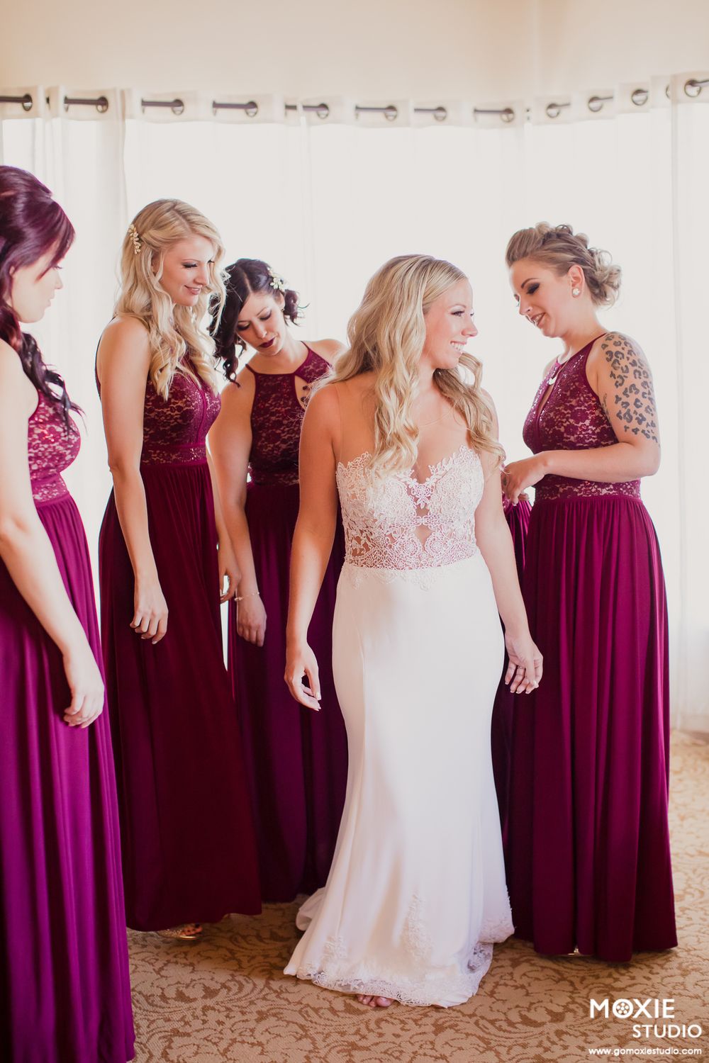 Bridesmaids in burgundy floor-length dresses share a candid moment with bride in white lace gown.