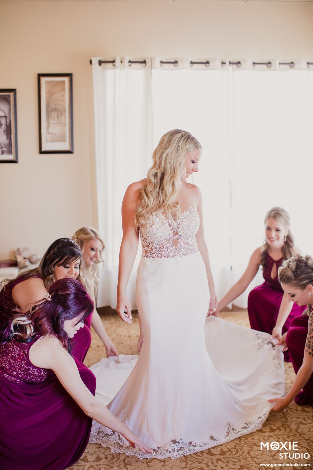 A wedding party in purple bridesmaid dresses helps adjust the bride's white gown in a sunlit room.