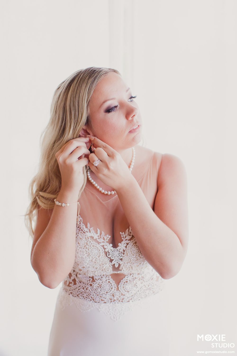 A bride in a white lace gown puts on pearl earrings while getting ready for her wedding day.