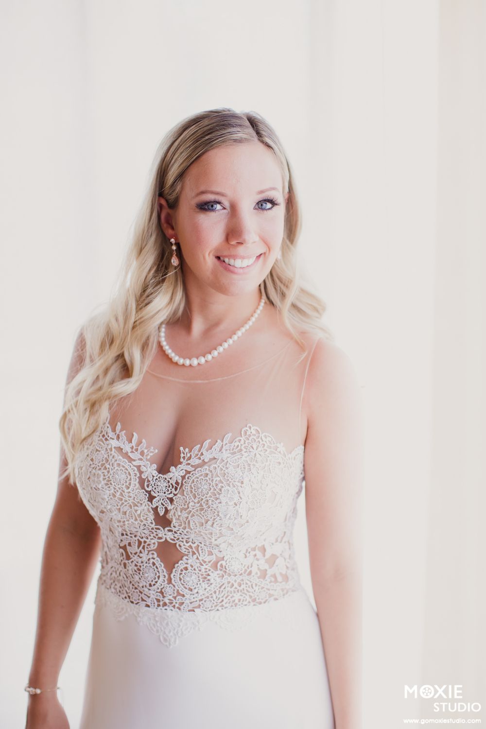A bride in a white lace wedding dress with intricate details and pearl necklace poses for bridal portraits.