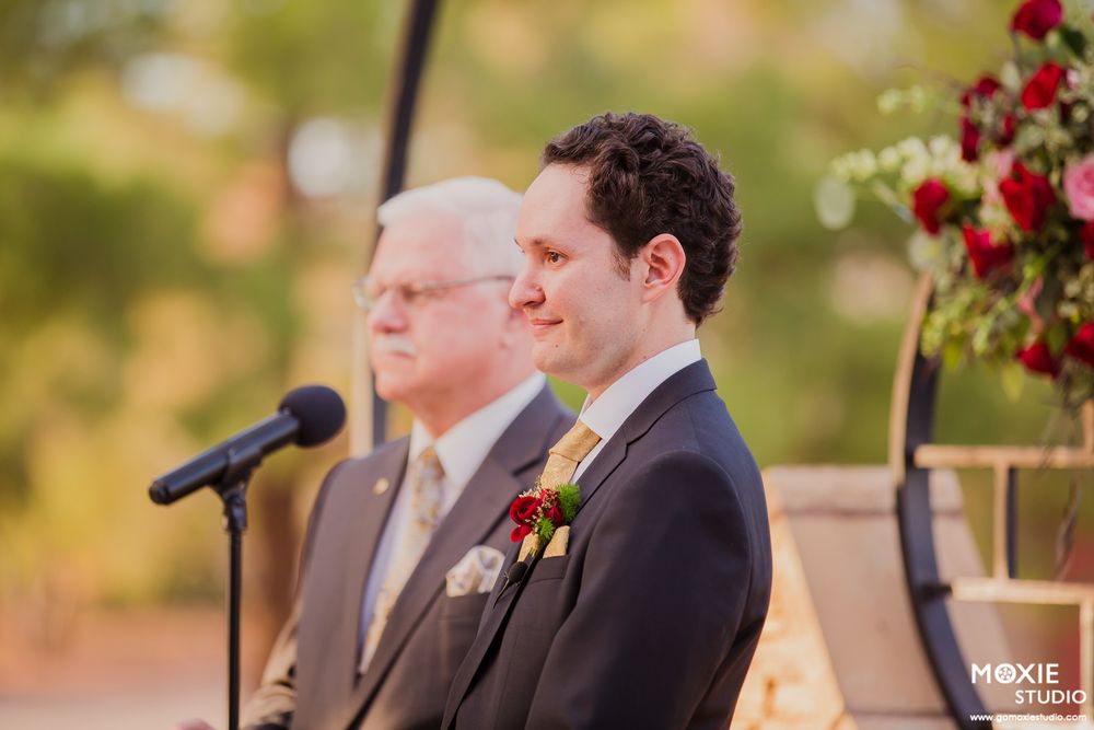 Two people stand together during an outdoor wedding ceremony with red floral decorations in the background.