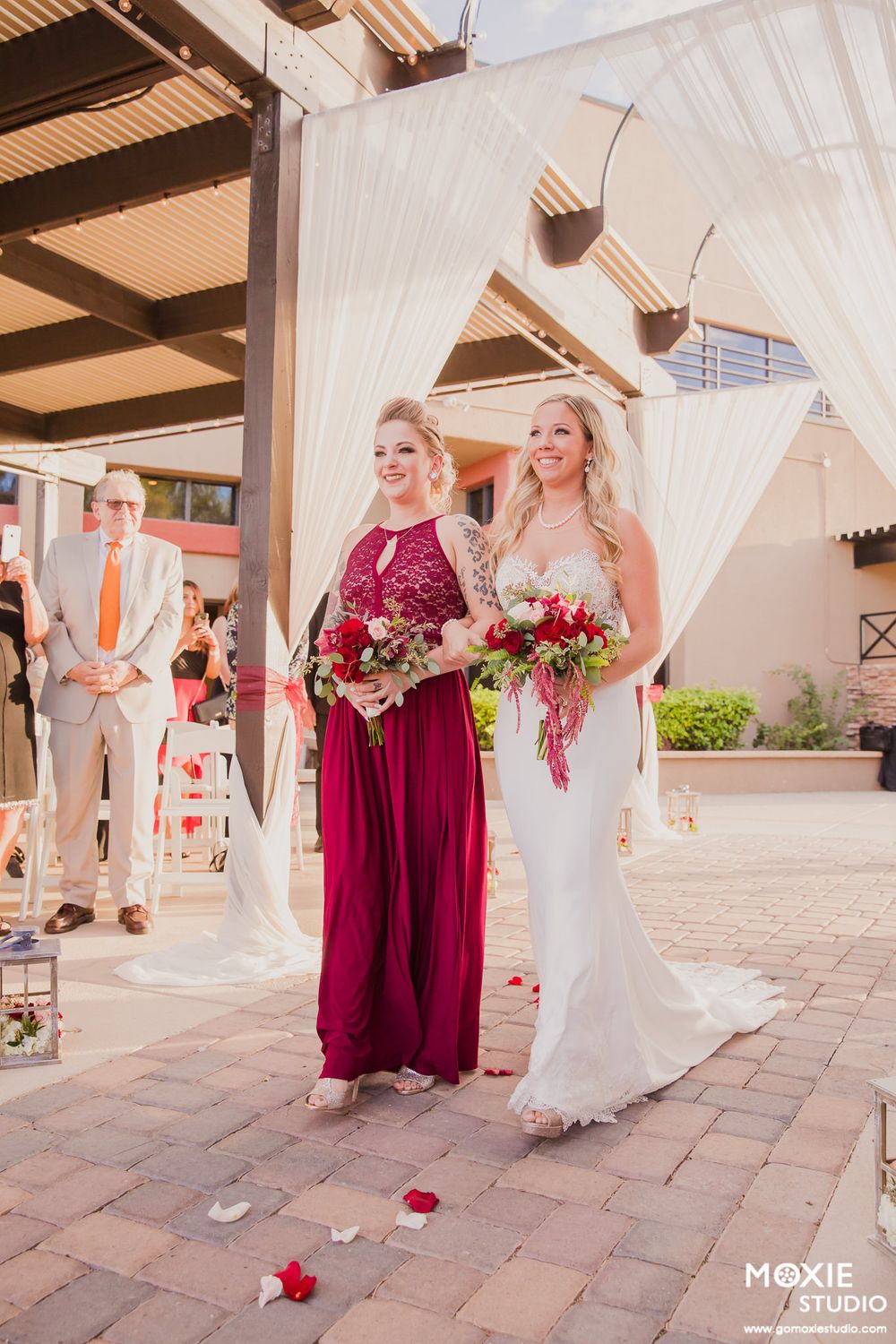 A wedding party in burgundy and white dresses poses on a paved courtyard with white curtains and wooden beams overhead.