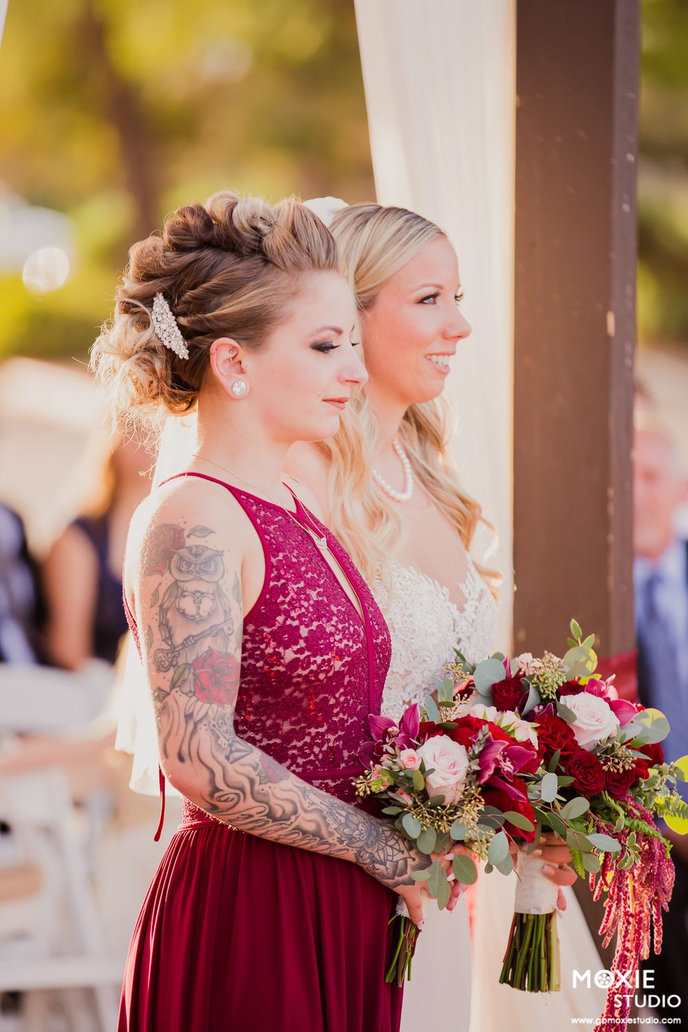 Two people in formal attire at a wedding ceremony, one in burgundy lace and one in white, holding floral bouquets.