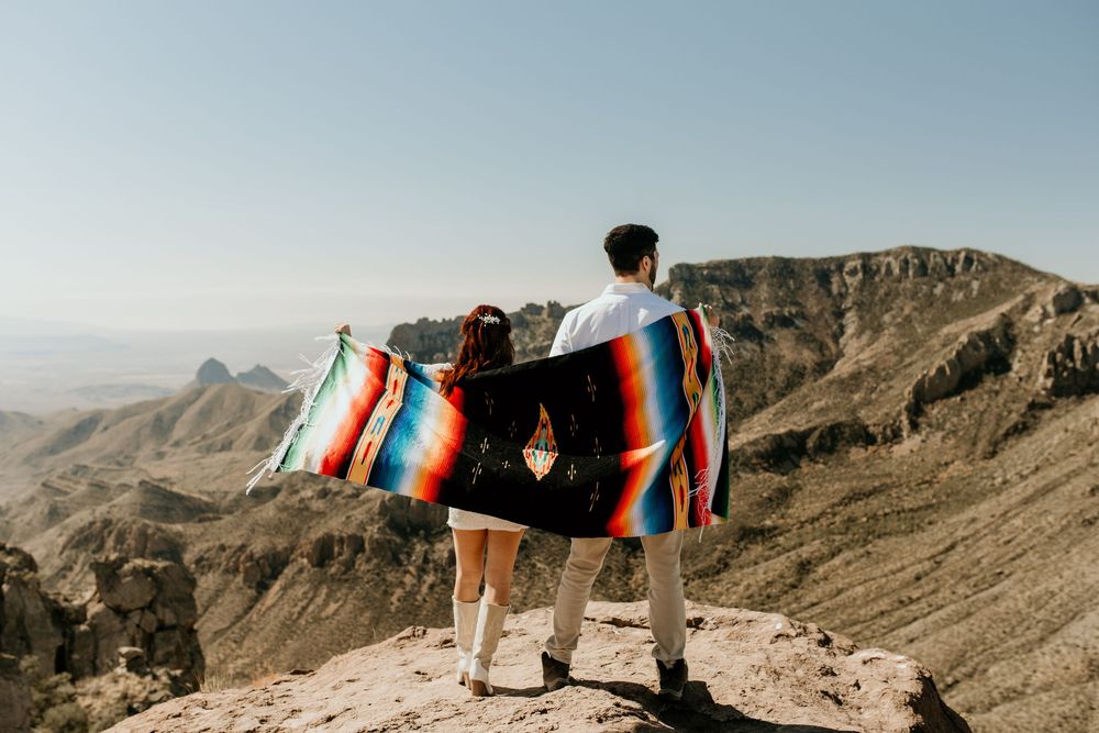 Two people wrapped in colorful blanket standing on mountain overlook with desert vista.