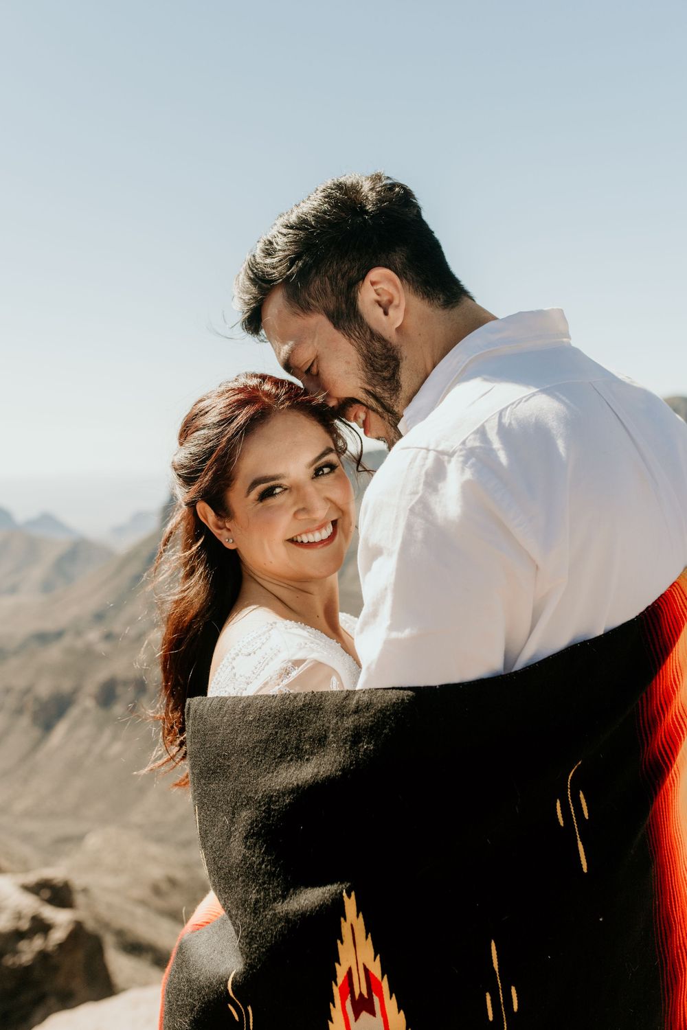 Couple sharing tender moment while wrapped in southwestern pattern blanket against mountain backdrop.