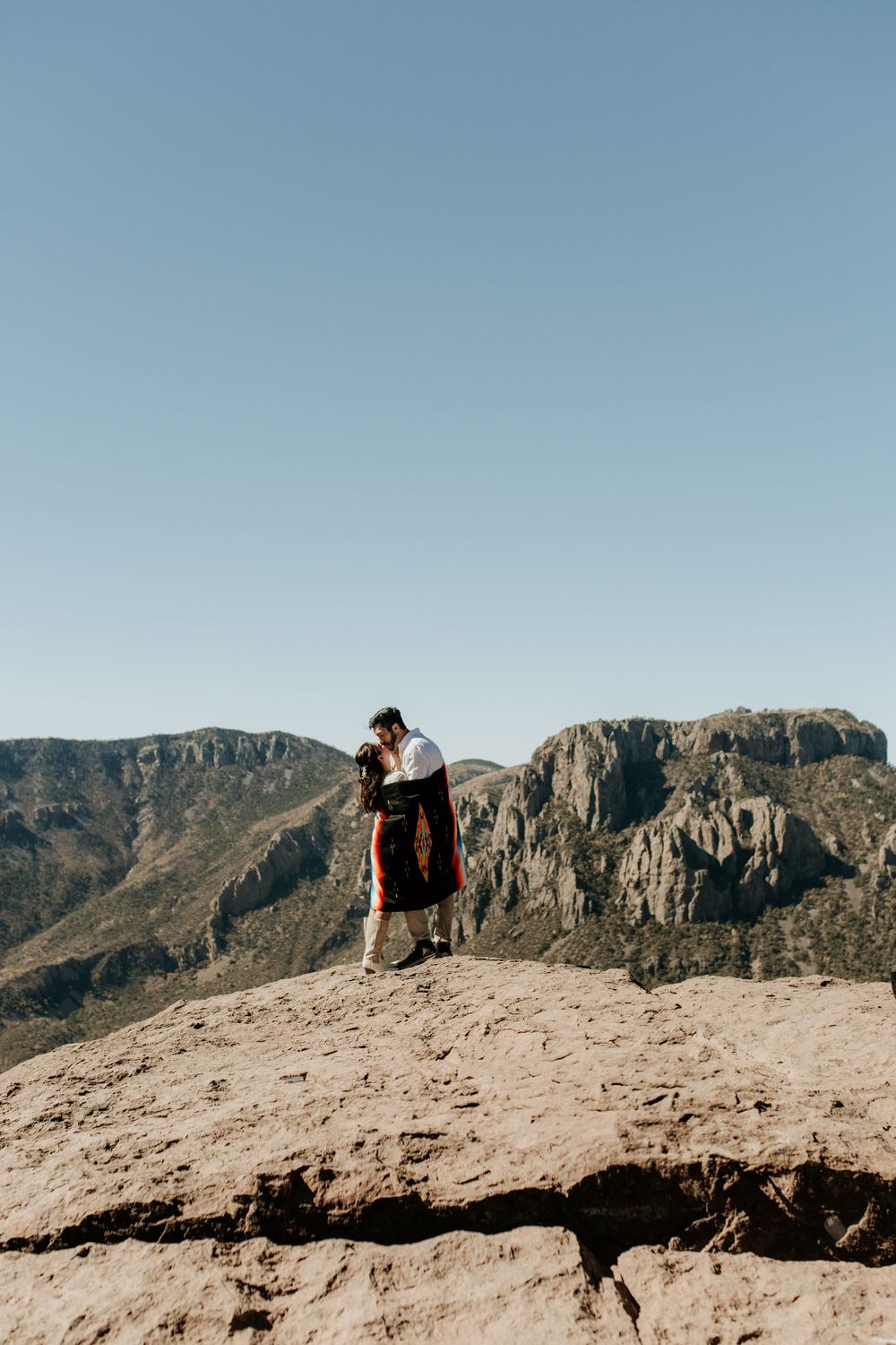 Couple standing together on desert mountain peak with dramatic cliffs in background.
